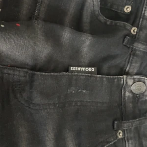 Svarta jeans från Dsquared2 - Snygga svarta jeans från Dsquared2 med slitna detaljer och färgstänk. De har en klassisk femficksdesign och knappgylf. Perfekta för en trendig look.