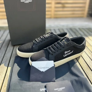  Saint Laurent skor - Snygga svarta sneakers från Saint Laurent med vit sula och klassisk snörning. Skorna har en stilren design med Saint Laurent-logga på sidan och tungan. Perfekta för en trendig och avslappnad look.