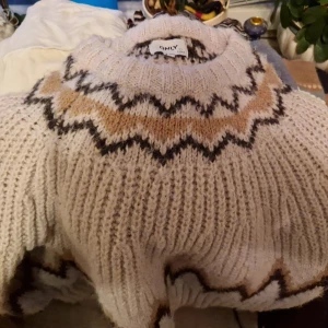 Vit stickad tröja från ONLY - Mysig vit stickad tröja från ONLY med ett snyggt mönster i svart och beige runt halsen och ärmsluten. Perfekt för kyliga dagar. Tröjan har en ribbad struktur och långa ärmar.