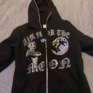 Svart hoodie med glittrig text - Snygg svart hoodie med dragkedja och glittrig text på framsidan. Designen inkluderar en måne och en figur, vilket ger en unik look. Perfekt för att sticka ut i mängden.