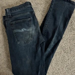 Mörkblå jeans från Nudie - Snygga mörkblå jeans från Nudie med klassisk femficksdesign och broderad bakficka. Perfekta för en avslappnad stil med en touch av elegans. Passar till alla tillfällen! Sällsynta