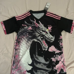 Svart och rosa Japan FIFA fotbollströja - Cool svart fotbollströja från Adidas med rosa detaljer och ett stort drakmotiv. Tröjan har korta ärmar och ett unikt tryck med japanskt tema, inklusive körsbärsblommor och traditionell arkitektur. Perfekt för fotbollsfans som vill sticka ut!