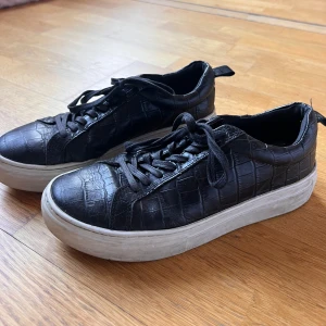 Svarta sneakers med krokodilmönster - Snygga svarta sneakers med ett coolt krokodilmönster. Skorna har en tjock vit sula och snörning framtill. Det är några skador på de, baksidan där flärpen har gett skador och ett hål i lädret på ena sidan av skon. Jag skulle inte säga att detta är något som syns annars eller som stör passformen.🌸