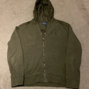 Grön hoodie från Polo Ralph Lauren - Säljer en snygg grön hoodie från Polo Ralph Lauren med dragkedja och huva. Perfekt för en avslappnad stil. Den har långa ärmar och en diskret logga på bröstet. Passar bra till det mesta, speciellt jeans eller träningsbyxor.