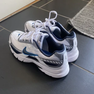 Nike sneakers i vitt och blått - Snygga Nike sneakers i vitt med blå detaljer och snörning. Perfekta för en sportig look med en bekväm passform. Den klassiska Nike-loggan syns på sidan och tungan av skon. 