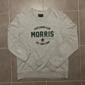 Morris Sweatshirt Grå M - Säljer en stilren grå sweatshirt från Morris med texten 'Gentlemens Club' och 'Est. Since 1968' i grönt på framsidan. Tröjan har långa ärmar och ribbade muddar vid ärmslut och nederkant. Perfekt för en avslappnad stil.