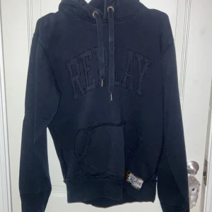 Svart hoodie från Replay - Säljer en svart hoodie från Replay med broderad logga på bröstet. Den har en klassisk känguruficka och justerbar huva med snören. Perfekt för en avslappnad stil.