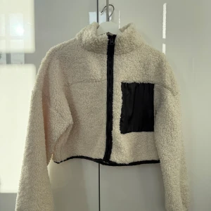 Beige teddyjacka med dragkedja - Säljer en mysig beige teddyjacka med dragkedja och en svart ficka på bröstet. Jackan är croppad och har långa ärmar, perfekt för kyliga dagar. 