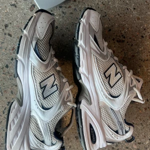 Vita och grå sneakers från New Balance - Snygga och bekväma sneakers från New Balance i vitt och grått med svarta detaljer. Skorna har en klassisk design med snörning och en stabil sula, perfekt för både vardag och träning.