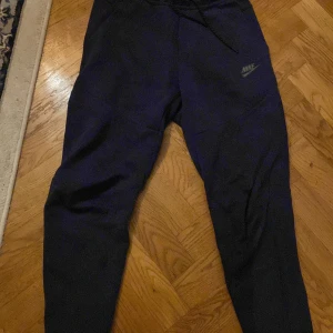 Blå mjukisbyxor från Nike - Säljer ett par bekväma blå mjukisbyxor från Nike med dragsko i midjan och praktiska fickor med dragkedja. Perfekta för en avslappnad stil eller träning. Nike-loggan är tryckt på benet för en sportig look.