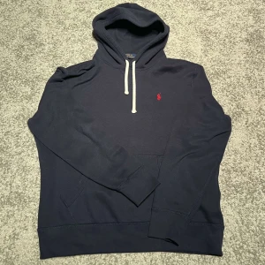Mörkblå hoodie från Polo Ralph Lauren - Säljer en snygg mörkblå hoodie från Polo Ralph Lauren med en röd broderad logga på bröstet. Tröjan har en justerbar huva med vita snören och en praktisk magficka. Perfekt för en avslappnad stil.