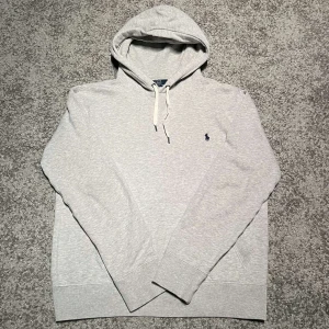 Grå hoodie från Ralph Lauren - Säljer en stilren grå hoodie från Ralph Lauren med broderad logga på bröstet. Tröjan har en justerbar huva med snören och långa ärmar. Perfekt för en avslappnad look.