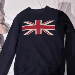Mörkblå stickad tröja med Union Jack från Crocker - Säljer en mörkblå stickad tröja från Crocker med Union Jack-mönster på bröstet. Tröjan har långa ärmar och ribbstickade muddar vid ärmslut och nederkant. Perfekt för en stilren och avslappnad look. Passar både män och kvinnor