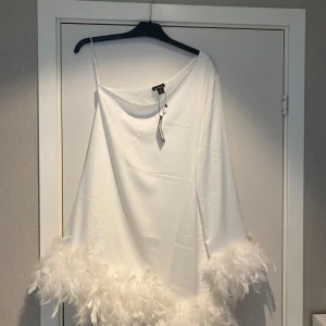 Vit klänning med fjäderdetaljer - Elegant vit, enarmad klänning från Bohoo med fjäderdetaljer längs fållen. Perfekt för speciella tillfällen som t.ex student/skolavslutning. Klänningen har en normal passform, storlek 38. Ny, Aldrig använd.  