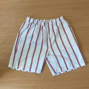 Randiga shorts - Säljer ett par vita shorts med röda och blå ränder. De har en elastisk midja för extra komfort. Perfekta för en avslappnad sommarstil.