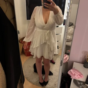 Vit omlottklänning med volanger från Boohoo - Supersöt vit kortklänning från Boohoo med omlottmodell, volanger och knyt i midjan. Klänningen har långärmade, lätt transparenta ärmar och djup v-ringning. Perfekt till student eller skolavslutning☀️🎉Ej använd prislapp kvar