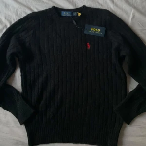 Svart stickad tröja  Polo Ralph Lauren - Kabelstickad svart tröja från Polo Ralph Lauren med rund halsringning och röd broderad logga på bröstet. Klassisk och stilren modell med långa ärmar och ribbade muddar. Perfekt för lager-på-lager.