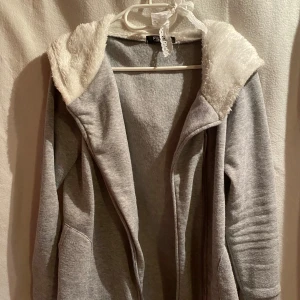 Grå hoodiejacka med fluffig huva - Mysig grå hoodiejacka från Fishbone med stor, fluffig vit huva i fuskpäls. Jackan har långa ärmar och dragkedja framtill. Perfekt för kyliga dagar när du vill vara både bekväm och snygg.