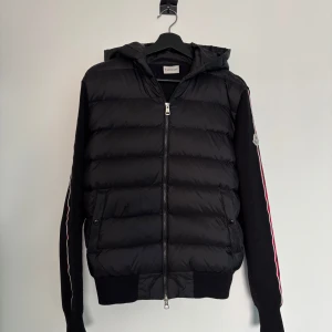Cardigan från Moncler - Snygg svart cardigan från Moncler med huva och dragkedja. Jackan har långa ärmar med röd och vit randdetalj samt Moncler-logga på ärmen. Storlek M men är kort i midjan.