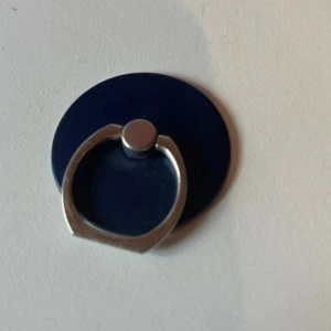 Mörkblå mobilring - Säljer en stilren mörkblå mobilring med rund form och silverfärgad ring. Perfekt för att få bättre grepp om mobilen eller använda som ställ. Enkel och snygg design som passar de flesta mobiler. Helt oanvänd med klistermärket kvar där bak för limmets skull