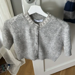 Grå kofta med pärl- och strassdetaljer från Zara - Superfin grå kofta från Zara med kort, croppad passform. Koftan har pärl- och strassdetaljer runt kragen och knäppning framtill. Mjuk och elegant på samma gång. Ordinarie pris 299kr, storlek M