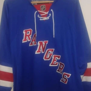 New York Rangers autentisk Jeresey från 2005 - Säljer en blå hockeytröja från Reebok med Rangers-logga på framsidan. Tröjan har långa ärmar med vita och röda ränder samt snörning vid halsen. Perfekt för hockeyfans! 🏒