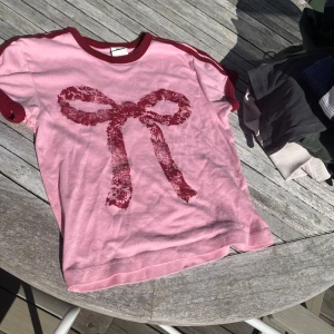 Rosa t-shirt med rosett från Urban Outfitters - Säljer en rosa t-shirt från Urban Outfitters med vinröda kanter och ett stort rosettmotiv framtill. T-shirten har korta ärmar och är i mjuk bomull, perfekt för en avslappnad stil.