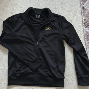 Svart track jacket från Emporio Armani EA7 - Säljer en svart track jacket från Emporio Armani EA7 med en guld logga på bröstet och dekorativa ränder på ryggen. Jackan har dragkedja framtill, ståkrage och ribbade muddar. Perfekt för en sportig och stilren look.