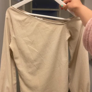 Beige långärmad topp med bred halsringning - Säljer en beige långärmad topp med bred halsringning. Toppen har en lätt ribbad struktur och är perfekt för en stilren och enkel look. Passar dig som gillar neutrala färger och en avslappnad passform.