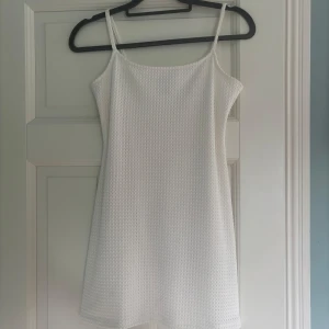 Vit ärmlös klänning från H&M Divided - Säljer en vit kortklänning från Divided med smala axelband och diskret strukturmönster i tyget. Klänningen har en figursydd passform och är tillverkad i ett lätt material, perfekt för varma dagar. Använd 1 gång vid skolavslutning.