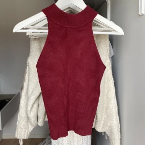 Vinröd ribbad ärmlös topp - Säljer en vinröd ribbad topp med hög hals och ärmlös design. Toppen är figurnära och har en mjuk, stretchig känsla. Perfekt att styla med jeans eller kjol för en trendig look. Stretchigt material.