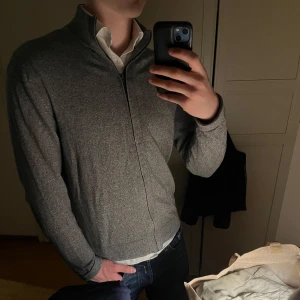 Grå Kashmir tröja  från Tommy Hilfiger - Snygg grå tröja från Tommy Hilfiger i en mjuk bomulls- och kashmirblandning. Tröjan har en praktisk dragkedja framtill och en stilren design som passar perfekt för kyligare dagar.