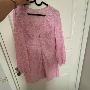 Rosa mönstrad klänning från Nelly x Glamorous - Säljer en söt rosa klänning med ett diskret mönster från Nelly x Glamorous. Klänningen har långa ärmar och knappar framtill, perfekt för en somrig look. Den är i storlek 34 och har en v-ringad design.