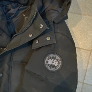 Svart dunjacka från Canada Goose - Säljer en svart dunjacka från Canada Goose med huva och diskret logotyp på ärmen. Jackan har dragkedja och knappar framtill samt stora fickor. Perfekt för kalla dagar när du vill hålla dig varm och bekväm.