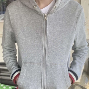 Grå hoodie med dragkedja från Moncler - Säljer en stilren grå hoodie från Moncler med dragkedja och huva. Jackan har ribbade muddar med röd, vit och blå detalj samt Moncler-logga på ärmen. Perfekt för en avslappnad look. vid fler funderingar eller bilder är det bara att höra av er.