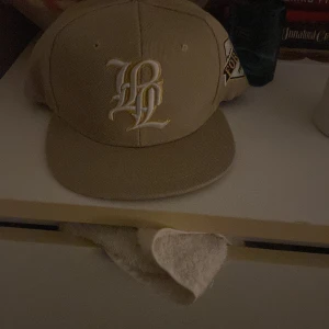 Beige keps med broderad logga och patch - Snygg beige keps med vit och gul broderad logga framtill och en patch på sidan med texten 'FOREVER PHASE-001'. Justerbar baktill för perfekt passform. Perfekt accessoar för en avslappnad stil.