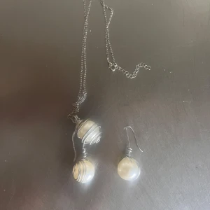 Smyckesset med halsband och örhänge med pärldetaljer - Säljer ett set med halsband och ett örhänge i silverfärgad metall med stora vita pärlor inlindade i tråd. Kedjan är tunn och smyckena har en modern, handgjord känsla. Perfekt för dig som gillar unika accessoarer.