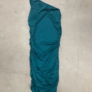 Turkos one shoulder långklänning - Säljer en turkos långklänning med one shoulder-design och rynkade detaljer längs sidan. Klänningen har en smickrande passform och är tillverkad i ett mjukt, stretchigt material som känns bekvämt mot huden.
