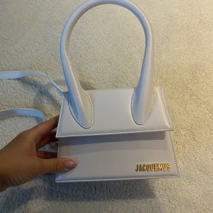 Äkta handväska från Jacquemus - Snygg vit handväska från Jacquemus med ett stilrent och modernt utseende. Väskan har en justerbar axelrem och ett elegant guldfärgat spänne. Perfekt för att ge din outfit en lyxig touch.