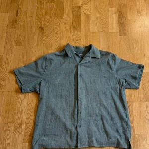 Grågrön kortärmad skjorta med knappar - Säljer en stilren grågrön skjorta med korta ärmar och klassisk krage. Skjortan har knäppning framtill och en avslappnad passform, perfekt för varmare dagar. Enkel och tidlös design som passar till många tillfällen.