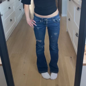 Blå bootcut jeans med slitningar - Säljer ett par blå bootcut jeans med slitna detaljer och låg midja. Jeansen har klassiska fickor fram och bak samt synliga sömmar. Perfekta för en avslappnad och trendig look.