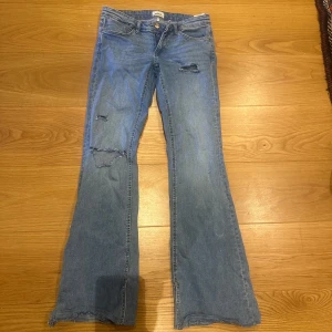 Blå bootcut jeans med slitningar - Säljer ett par blå bootcut jeans med slitningar på låret och knät. Jeansen har klassisk femficksdesign, låg midja och råa kanter vid bensluten. Perfekta för en avslappnad och trendig look. Dom är insydda då det var en slits innan 
