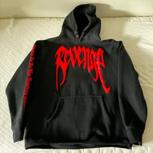 Svart hoodie från Revenge med röd text - Svart hoodie från Revenge med stor röd logga på bröstet och tryck på ena ärmen. Baksidan har ett stort rött handavtryck. Klassisk känguruficka och huva med metallöljetter.