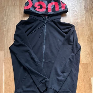 Svart hoodie med dragkedja från HUGO - Svart hoodie från HUGO med dragkedja och stor huva med röd logotyp. Tröjan har fickor framtill och ribbade muddar vid ärmslut och nederkant. Perfekt för en avslappnad stil.