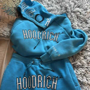 Blå Set från Hoodrich - Säljer en blå set med byxor och hoodie från Hoodrich i Storlek M med stor vit logga framtill och broderade detaljer på huvan och ärmen. Hoodien har dragsko i huvan och en magficka. Perfekt för dig som gillar streetwear och vill sticka ut. Använt byxorna 1 gång och hoodien inte många gånger alls. Självklart tvättas kläderna innan dem skickas ut. 