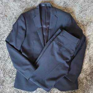 Marinblå kostym från Bläck (MQ) - Elegant marinblå kostym med kavaj och byxor. Kavajen har två knappar och klassiska slag. Byxorna är raka och stilrena. Bara använd 2 gånger och perfekt för formella tillfällen.