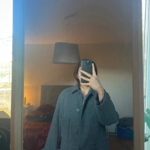 Grå kappa från 157 - Säljer en stilren grå ulljacka från 157. Jackan har en klassisk krage och knäppning med stora knappar framtill. Den har långa ärmar och praktiska fickor. Perfekt för kyligare dagar!