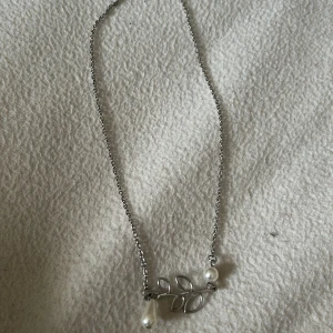 Silverfärgat halsband med pärlor - Elegant silverfärgat halsband med ett bladmotiv och två vita pärlor. Perfekt för att ge en stilren touch till din outfit.