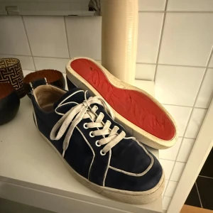 Christian Louboutin - Säljer ett par blå sneakers från Christian Louboutin med vit sula och vita snören. Skorna har röda sulor undertill, klassisk rund tå och kontrasterande vita detaljer längs kanterna. Perfekta för dig som vill sticka ut med exklusiva sneakers.
