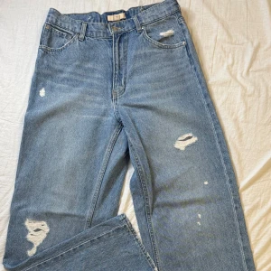Blå slitna jeans från H&M - Säljer ett par blå jeans från H&M med slitna detaljer och raka ben, hålen är lappade med lite mörkare jeans. Jeansen har klassisk femficksdesign, hög midja och är tillverkade i bomull. Perfekta för en avslappnad och trendig look. 